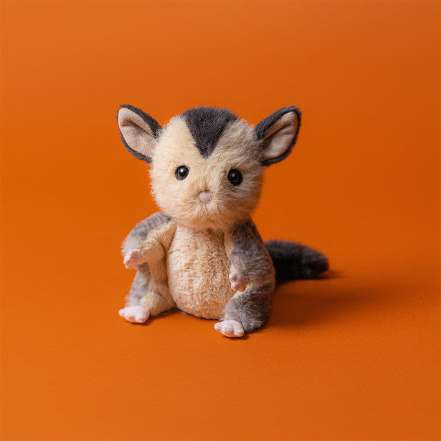The Jellycat Lolly Sugar Glider by Jellycat is a small, plush soft toy with large ears, a bushy tail, and brown-beige fur, sitting on an orange surface against an orange background.